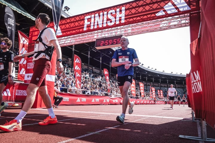 A 58-year-old runner, Mikael Bask, finishing the 2025 Stockholm Marathon under the red finish arch with a time of 2:59:56, watched by a crowd in the stadium.
