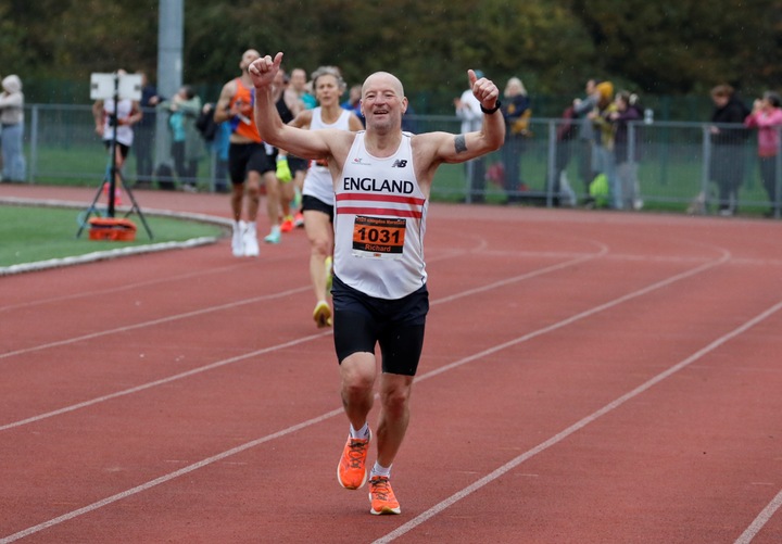 Richard Ginn running on the track in an England Masters vest, raising both arms as he approaches the finish.