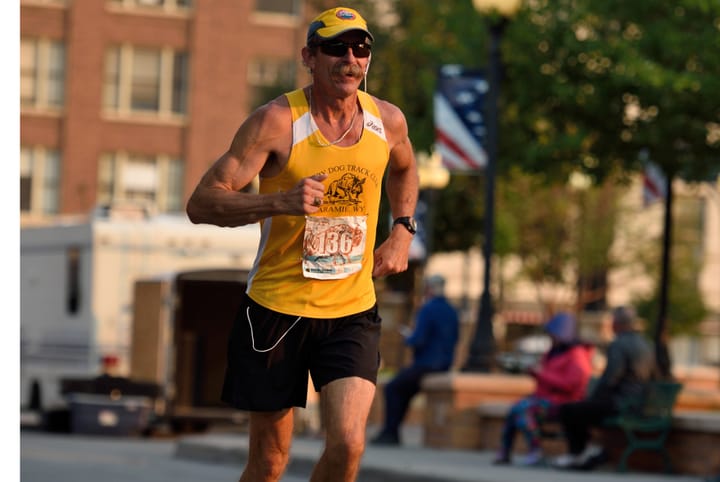Jerry Rief running a road race in a yellow Laramie, Wyoming track-club vest and cap, mid-stride on a city street with spectators and buildings blurred in the background.